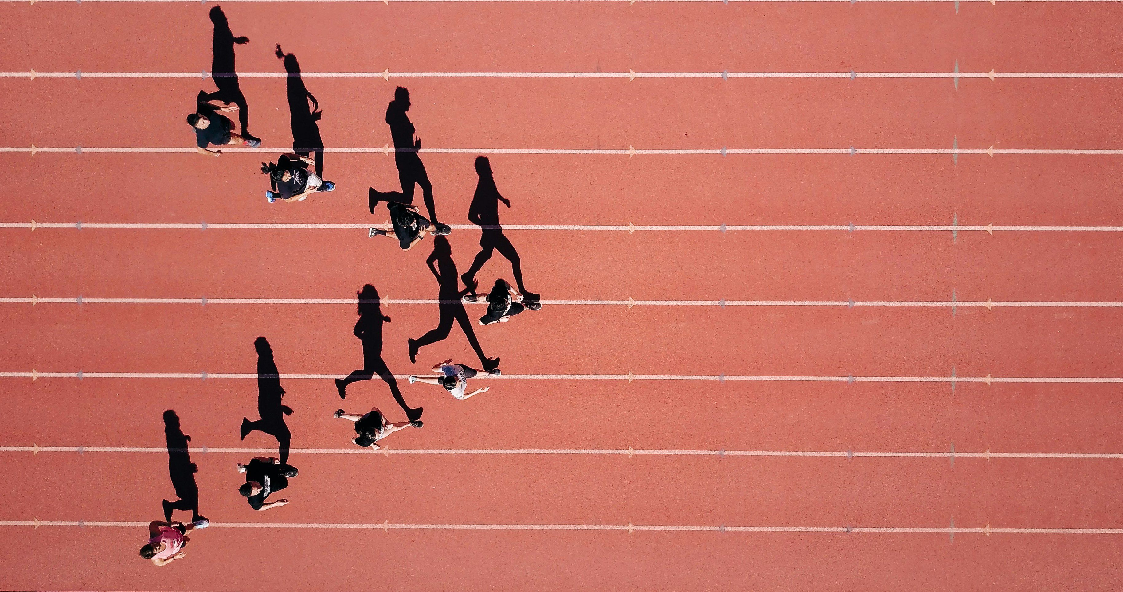 Aerial view of people running on a red track, with long shadows cast beside them in the sunlight.